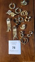 Photo showing multiple pairs of gold-tone clip-on earrings of various sizes and styles arranged on wood surface, including twisted hoops and dangling tassel designs.