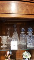 Full view of crystal lot showing all six pieces: two rectangular decanters with stoppers, one tall shaped decanter, one small rounded decanter, a footed glass vessel, and a crystal vase.