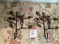 Pair of metal wall hooks shaped as branches with leaves and birds perched on them, dark metallic finish, displayed on a floral fabric background.