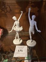 Photo of two white bisque porcelain dancer figurines, a male dancer and a ballerina, displayed on glass shelf with wooden background