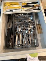 Top-down view of metal mesh drawer organizer containing assorted cutlery and knives, and a white plastic tray with various yellow-handled kitchen tools and black-handled basting brushes.
