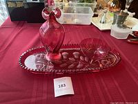 Photo showing a cranberry red crystal glass set: decanter with stopper, bowl, and etched oval tray on a red tablecloth.