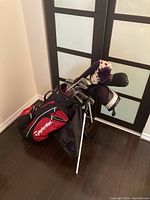 Golf club bag shown standing upright with golf clubs inside (clubs not included in sale). Shows bag's red and black design with TaylorMade branding and white stand legs.