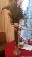 Full view of the tall clear glass vase holding multiple peacock feathers, tied with a brownish ribbon bow at the neck of the vase, placed on a table near a window.