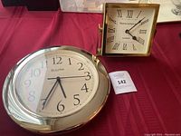 Photo of the two clocks: a round Bulova wall clock with silver frame and a square Thomas Sabo table clock with gold-tone frame.