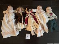 Five assorted vintage dolls laid on dark floor: two porcelain baby dolls in white christening gowns, one porcelain girl in brown velvet coat, one porcelain girl in red Victorian dress, one vinyl boy doll in knit sweater