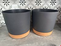 Two black cylindrical plant pots with terracotta-colored bases placed on a gray surface against a patterned wall.