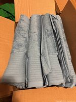 Four light blue cotton bath towels folded and aligned inside a cardboard box, showing texture and ribbed edges.