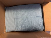 Single light blue towel folded inside a cardboard box showing the fabric texture and stripe detail.