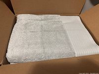 Four light gray 100% cotton bath towels neatly folded inside a cardboard box showing the texture and woven border detail.