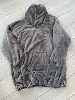Grey plush blanket sweater with hood laid flat on floor showing pocket and drawstring bottom