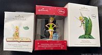 Three Hallmark Keepsake Tinkerbell Disney Christmas ornaments in their boxes arranged on a black background.