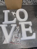 Photo of four white LED light-up letters spelling LOVE laid out with plastic mounting brackets and power cords still in packaging, showing all contents of the lot.