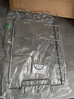 Metal pot lid holder with multiple vertical slots and three hooks at bottom, packaged in clear plastic. Mounting screws visible separately. Holder is silver metal with curved cutouts for lids.