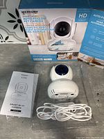 Full view of the pet camera on its plastic tray, showing the camera, white USB cable, and user manual next to the merchandise box.