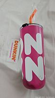 Full view of pink Dunkin Donuts stainless steel water bottle with large white letters, clear lid and orange straw wrapped in plastic.