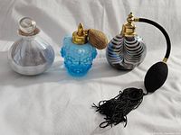 Three vintage-style perfume bottles displayed side by side on a white fabric background: silver-tone round bottle with glass stopper, blue hobnail bottle with gold atomizer and bulbed sprayer, and multicolored striped bottle with gold atomizer bulb and black tassel.