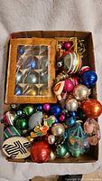 Full view of box containing assorted vintage glass Christmas ornaments in various colors and sizes including round baubles, bird figurine, and boxed sets.