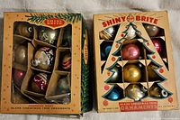 Two original Shiny Brite boxes containing vintage glass Christmas tree ornaments separated by cardboard dividers. Boxes show manufacturer logos and product information.