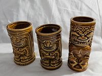Three brown ceramic tiki mugs with distinct tiki faces and poses, shown standing upright in a line