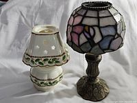 Pair of mini tealight lamps side by side on a white cloth background, showing stained glass style lamp with metal base on the right, and ceramic white floral lamp on the left.