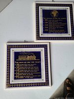 Both tiles shown side by side on a light floor. Each tile has intricate blue and white border design with gilt accents. The 'Woman is Like the World' tile has a globe image and text describing the woman humorously. The 'Man is Like the Train' tile features a steam engine train image and text.