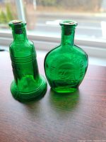 Two green glass miniature bottles with cork stoppers shown side by side on a wooden surface with a window in the background. The left bottle is shaped like the Liberty Bell. The right bottle is round with Benjamin Franklin's embossed face.