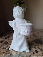 Front view of white unglazed ceramic angel holding a candle holder with detailed robe and wings.