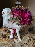 Side view of white ceramic angel holding flower pot with deep magenta silk roses and beige dried flowers on marble surface.