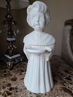 Front view of white, unglazed ceramic figurine of choir boy holding a bowl, standing on marble surface with decorative lamp in background.
