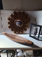 Sunburst mirror, carved wooden bowl, and framed African mask displayed together on a white surface with neutral background.
