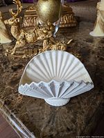 Front view of the white ceramic clam shell fan shaped holder on a marble surface with gold décor in the background, showing detailed ridges and shape.