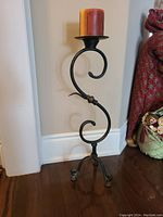 Front angle of tall hand forged S shape wrought iron candle holder with a red pillar candle on top, placed on wooden floor against white wall.