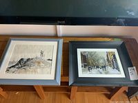 Two framed artworks on a wooden bench under a television: the left is a pen and ink landscape sketch with some brown ink wash, the right is a colored print of a snowy city street scene.