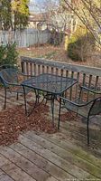 Black metal bistro set consisting of square table and two chairs on wooden deck outdoors.