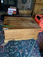 Wooden blanket box shown closed with two hinged lids and curved base detail, placed on a floral patterned rug next to other household items.