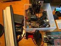 Overview of plastic bin containing vintage cameras and accessories including Kodak Instamatic, Polaroid Colorpak II, Praktica Super TL, small tripods, microphone with windscreen, and camera cases.