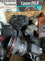 Close-up of woven basket filled with multiple Canon DSLR bodies and L-series red ring zoom lens.