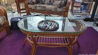 Side angled view of oval rattan coffee table with removable glass top and Bombay solitaire game set placed on top. Table on purple carpet with matching rattan furniture in background.