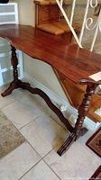 Console table viewed from the side showing scalloped edge on the top and turned legs connected by embossed crossbar.