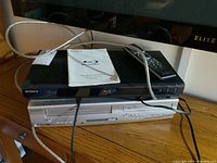 Sony black Blu-ray DVD player stacked on top of silver Sylvania VHS/DVD player on wooden surface. Visible cables and remote control on top of Sony player.