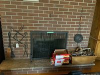 Photo of fireplace tools including broom, ash shovel, log lifter, and popcorn shaker arranged on the hearth against brick wall.