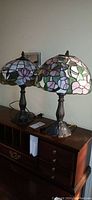 Two Tiffany style stained glass lamps on wooden dresser, side by side