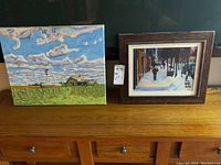 Photo showing both the oil on canvas painting of a rural landscape and the framed winter scene print side by side on a wooden surface.