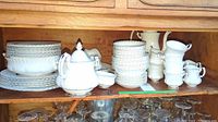 All pieces arranged on a wooden shelf showing a variety of stacked plates, bowls, teacups, and serving pieces in white with gold trim.