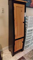 Full view of small wooden storage cupboard with natural wood doors and black painted frame and sides, tapered top design.