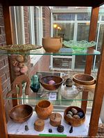 Photo showing wooden bowls, decorative eggs, carved figures, hydro insulator, oil lamp, and two glass plates in a glass cabinet.