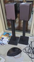 Photo showing two Monitor Audio bookshelf speakers on metal stands along with a curved black Mordaunt Short speaker on floor.