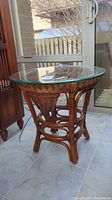 Full view of round rattan side table with loose glass top placed on tile floor near a windowed door.