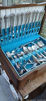Open canteen box displaying a full vintage cutlery set including knives, forks, tablespoons, teaspoons, and collector spoons with silver-tone ornate handles arranged on blue velvet lining.